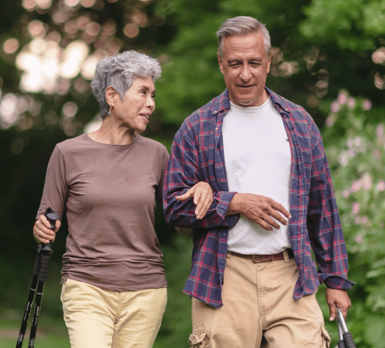 An elderly couple walks together outdoors, with the woman holding a walking stick and linking arms with the man who wears a plaid shirt, both smiling as they stroll through a green, tree-lined area.