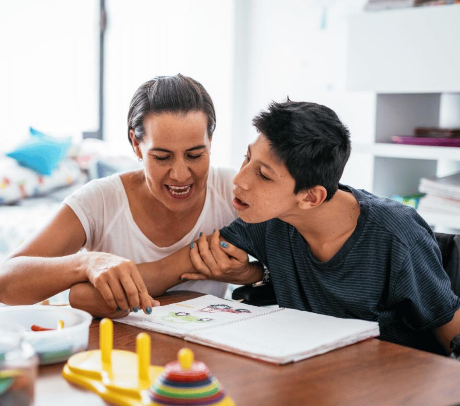 A woman and young boy sit together at a wooden table, smiling as they read from an open book, with colorful educational toys including stacking rings visible nearby in what appears to be a bright home learning environment.