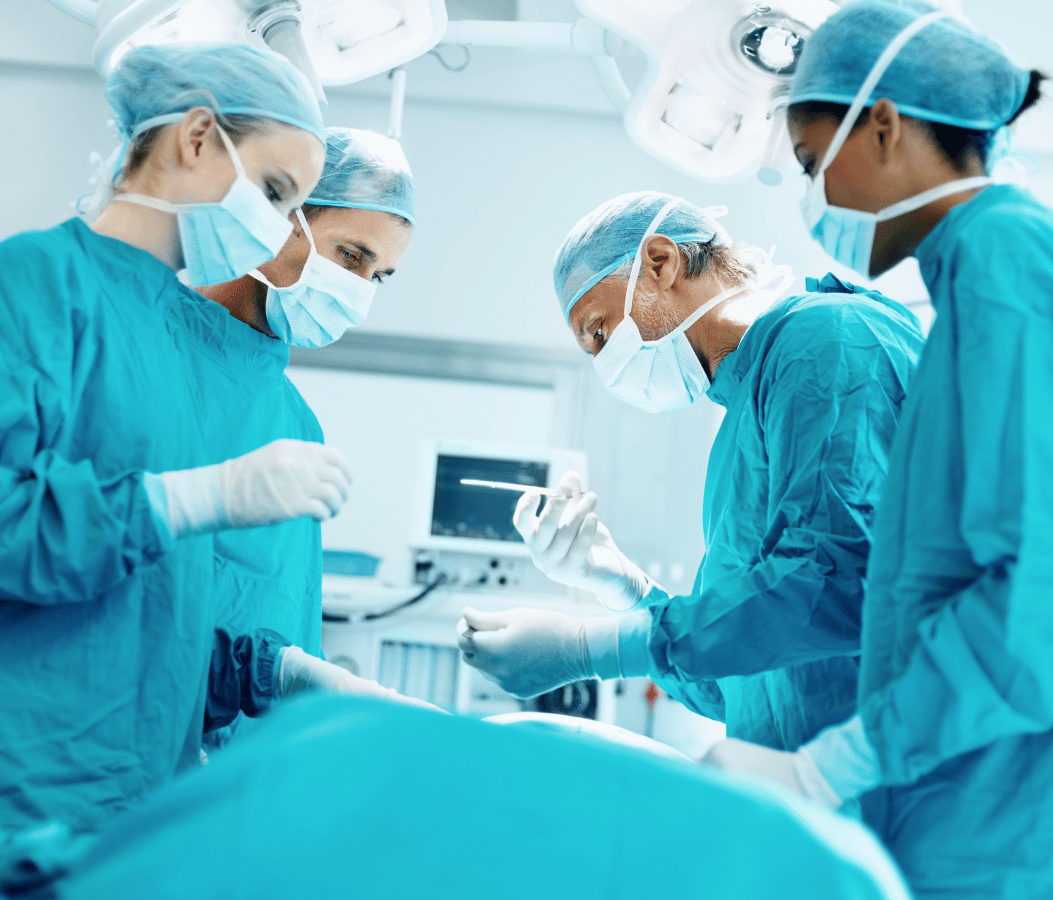 Four medical professionals in surgical scrubs, masks, and caps perform surgery in an operating room, working together around a patient under bright surgical lighting.