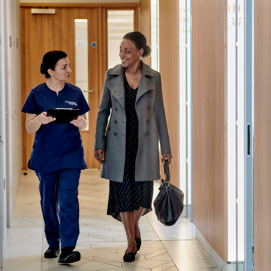 A healthcare worker in blue scrubs walks alongside a woman in a gray coat and striped dress through a modern hospital corridor, appearing to have a friendly conversation.