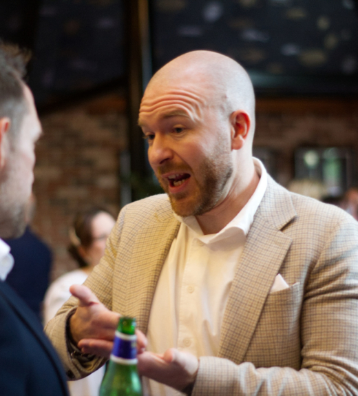 Two men in business attire having a conversation at what appears to be a networking event or business gathering, with one man in a light-colored suit jacket holding a green bottle while speaking to another man in a dark suit.