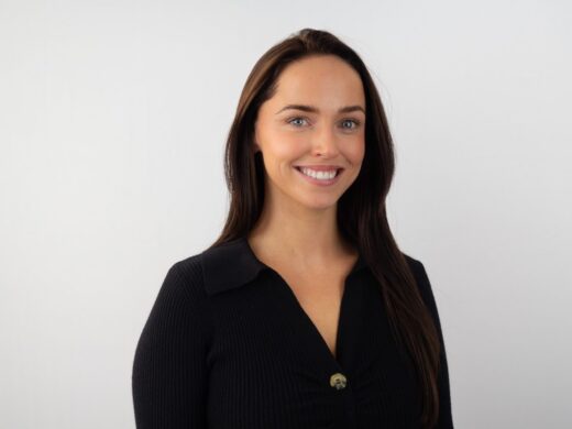 A woman with long dark hair smiles at the camera while wearing a black ribbed sweater with a collar and single button, photographed against a light gray background.