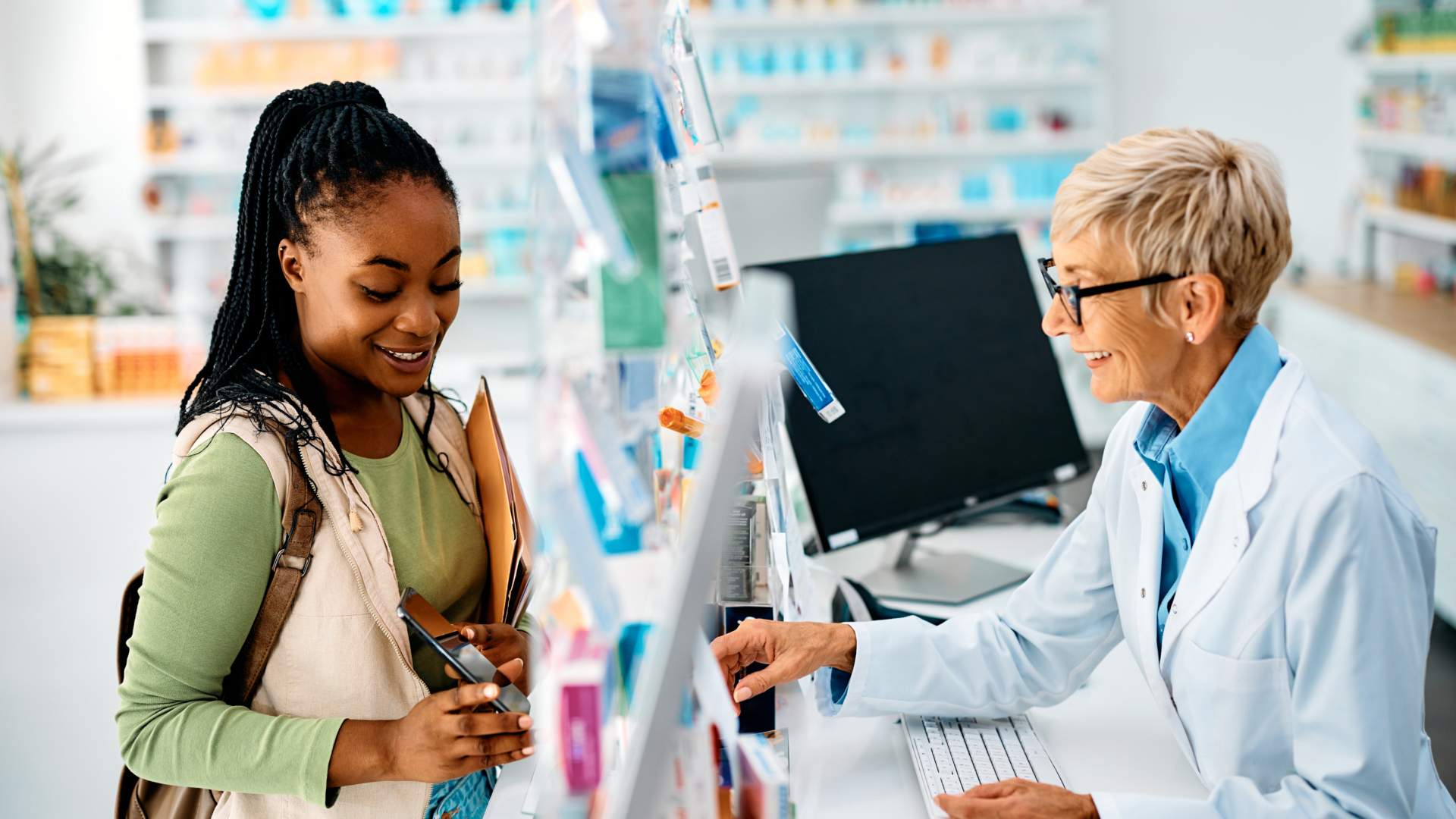 A young Black woman with braided hair speaks with an older white female pharmacist in a white coat at a pharmacy counter, with shelves of medications visible in the background.