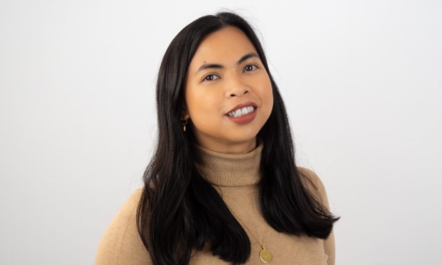 A woman with long dark hair wearing a beige turtleneck sweater and gold jewelry smiles warmly while looking slightly upward against a white background.
