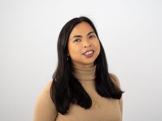 A woman with long dark hair wearing a beige turtleneck sweater and gold jewelry smiles warmly while looking slightly upward against a white background.