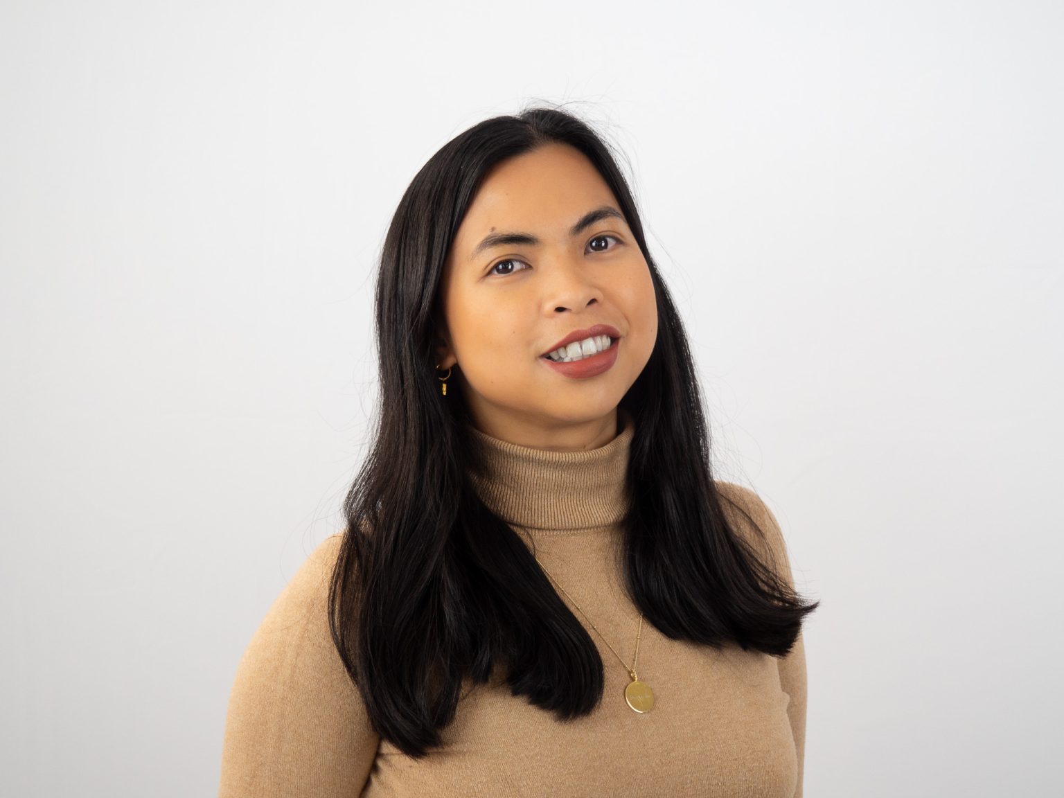 A woman with long dark hair wearing a beige turtleneck sweater and gold jewelry smiles warmly while looking slightly upward against a white background.