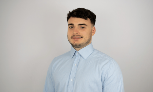 A young man with dark hair and facial hair smiles at the camera while wearing a light blue button-up shirt against a neutral background.