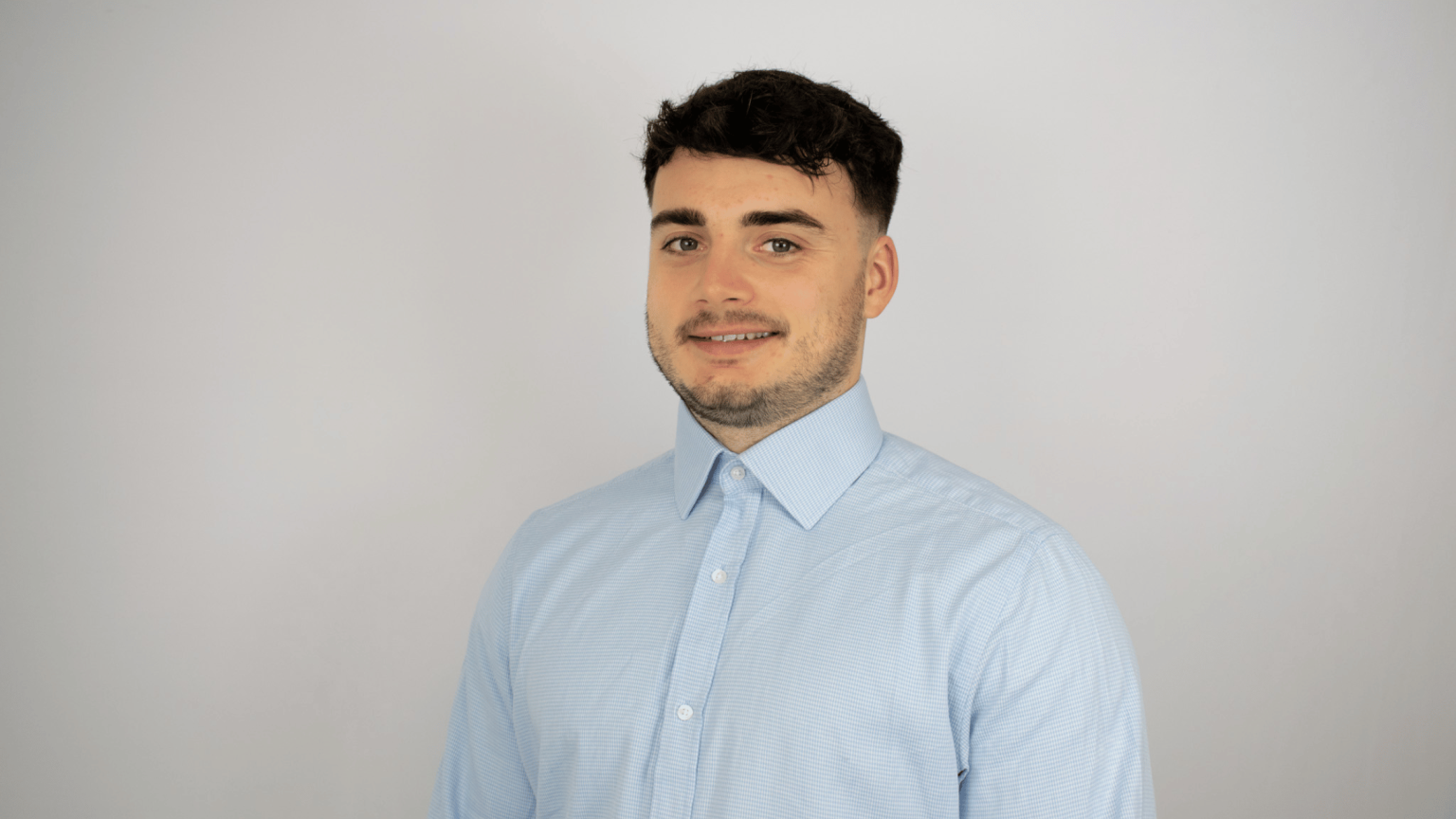 A young man with dark hair and facial hair smiles at the camera while wearing a light blue button-up shirt against a neutral background.