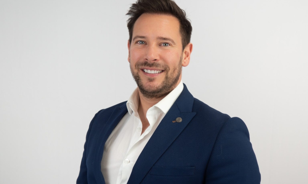 A professional headshot of a smiling man with dark hair and beard wearing a navy blue blazer over a white dress shirt against a light gray background.