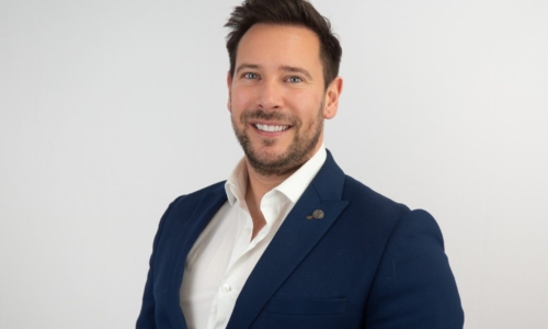 A professional headshot of a smiling man with dark hair and beard wearing a navy blue blazer over a white dress shirt against a light gray background.