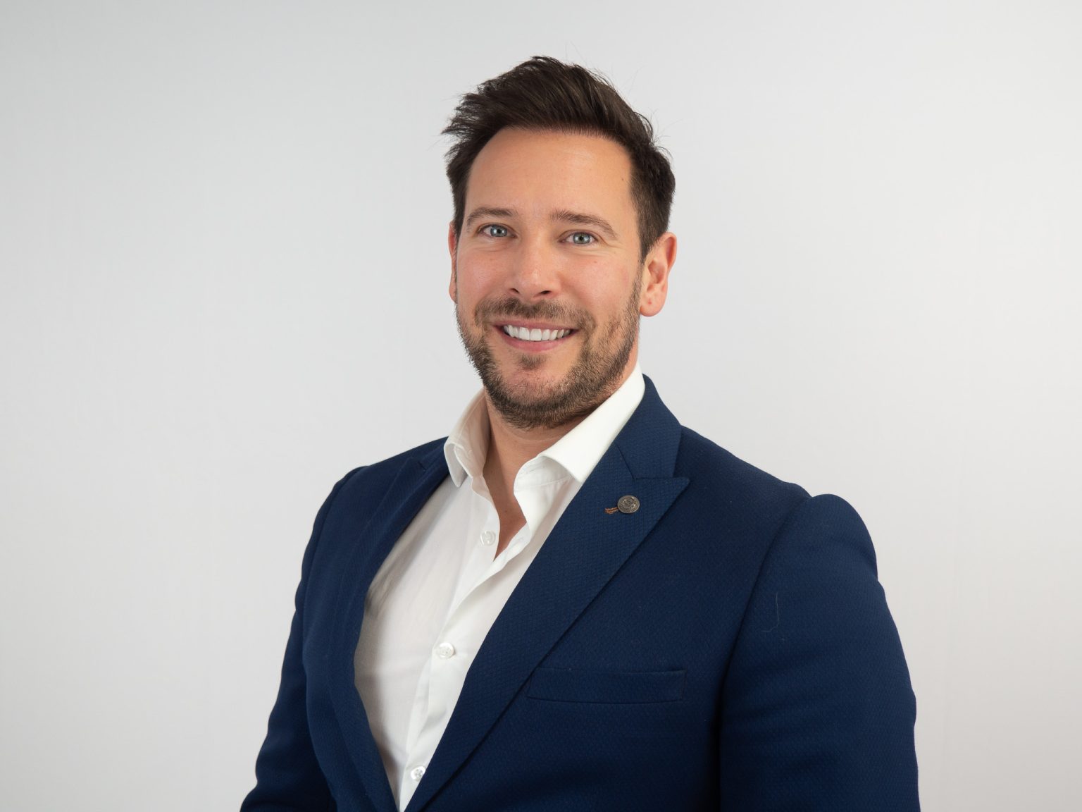 A professional headshot of a smiling man with dark hair and beard wearing a navy blue blazer over a white dress shirt against a light gray background.