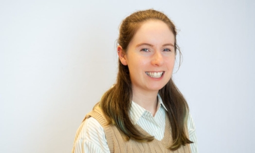 A young woman with long brown hair smiles warmly at the camera, wearing a beige cable-knit sweater vest over a light blue and white striped collared shirt against a white background.