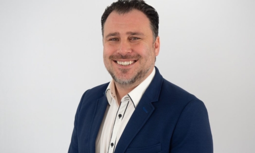 A smiling man with dark hair and facial stubble wearing a navy blue blazer over a white collared shirt, photographed against a plain white background in a professional headshot style.