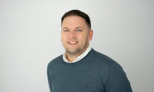 A man with short dark hair and facial stubble smiles at the camera, wearing a teal sweater over a white collared shirt against a plain gray background in what appears to be a professional headshot.