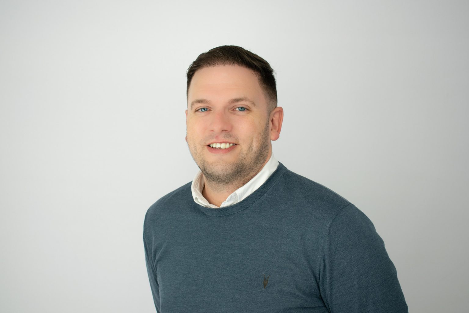 A man with short dark hair and facial stubble smiles at the camera, wearing a teal sweater over a white collared shirt against a plain gray background in what appears to be a professional headshot.