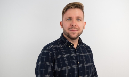 A man with short brown hair and a beard smiles at the camera while wearing a navy blue plaid button-down shirt against a white background.
