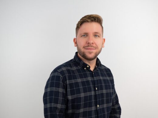 A man with short brown hair and a beard smiles at the camera while wearing a navy blue plaid button-down shirt against a white background.