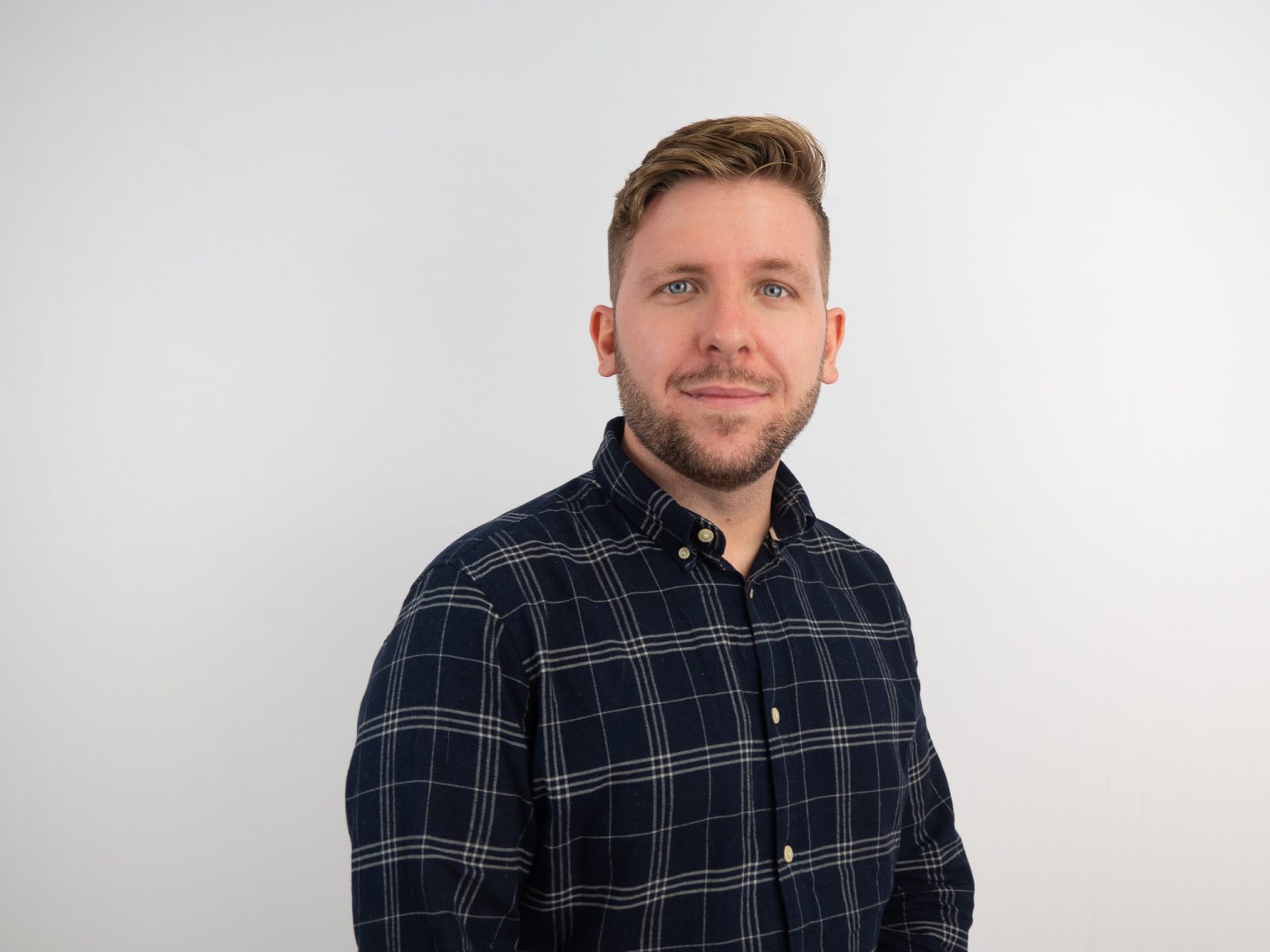 A man with short brown hair and a beard smiles at the camera while wearing a navy blue plaid button-down shirt against a white background.