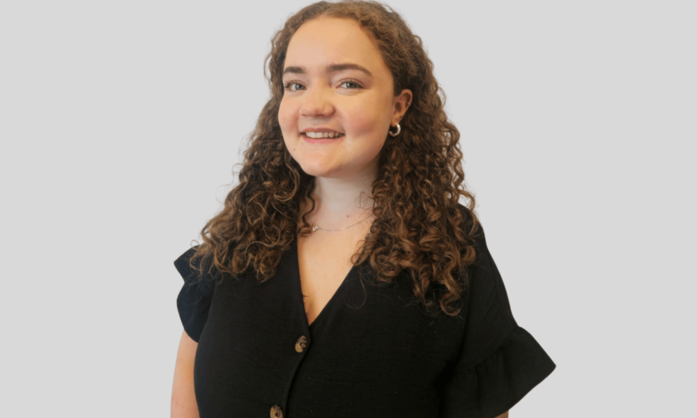 A young woman with curly brown hair smiles warmly while wearing a black button-up shirt with ruffled sleeves against a light gray background.