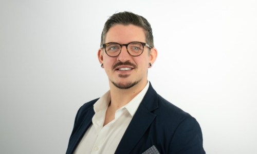 A professional man with glasses, a mustache, and ear piercings smiles at the camera while wearing a dark navy blazer over a white collared shirt with a gray pocket square.