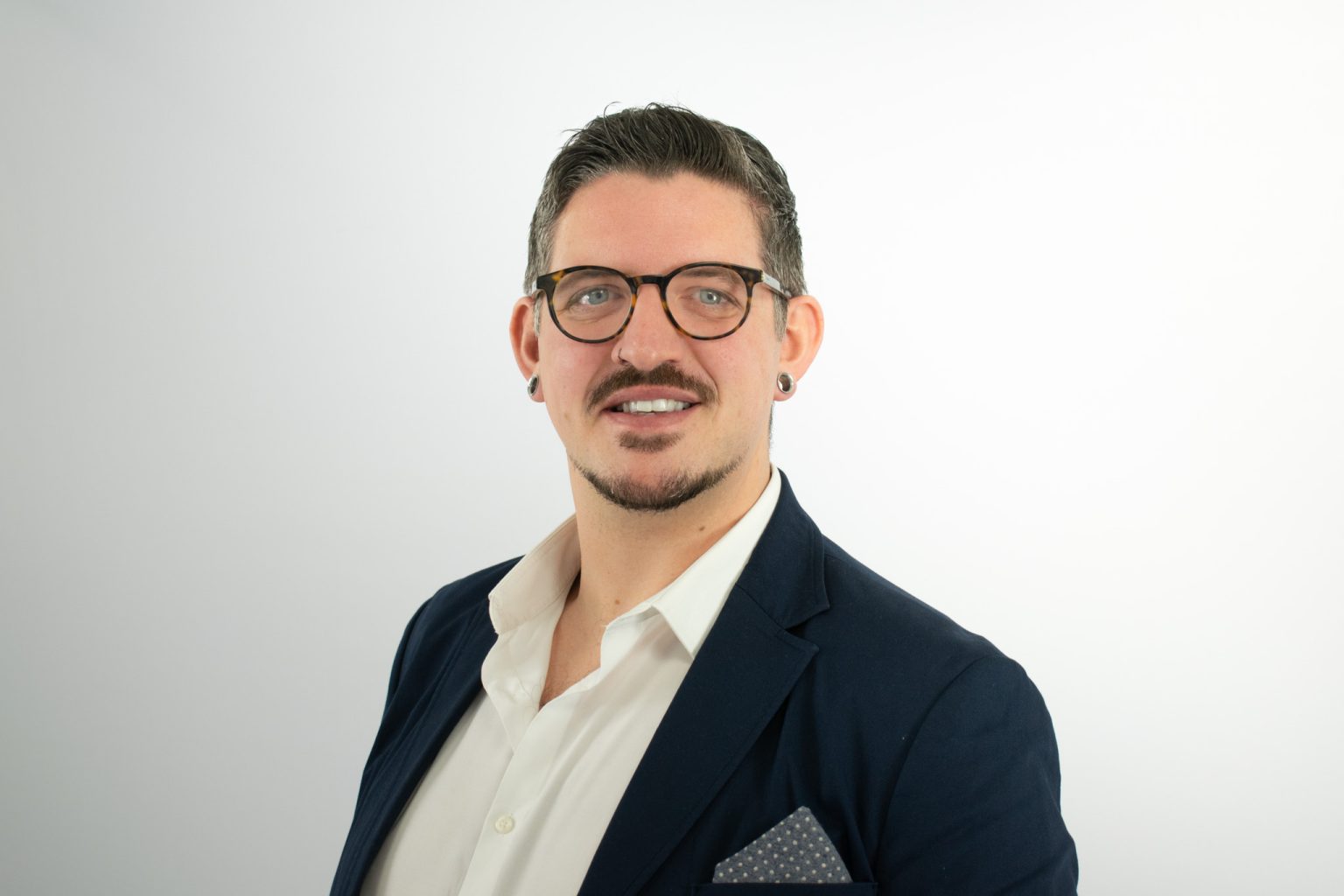 A professional man with glasses, a mustache, and ear piercings smiles at the camera while wearing a dark navy blazer over a white collared shirt with a gray pocket square.