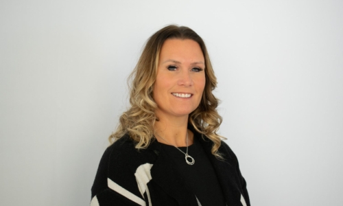 A professional woman with shoulder-length blonde wavy hair smiles at the camera, wearing a black sweater with white geometric patterns and a delicate necklace, photographed against a plain white background.