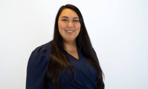 A woman with long dark hair smiles at the camera while wearing a navy blue wrap-style top, photographed against a white background in what appears to be a professional headshot.