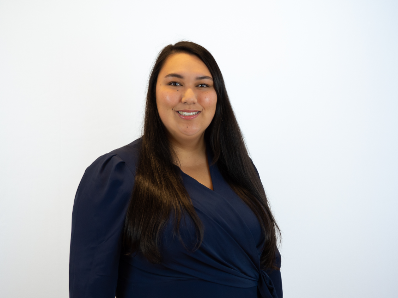 A woman with long dark hair smiles at the camera while wearing a navy blue wrap-style top, photographed against a white background in what appears to be a professional headshot.