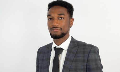 A professional headshot of a young Black man wearing a charcoal gray suit with a white dress shirt and black tie, smiling slightly at the camera against a light gray background.