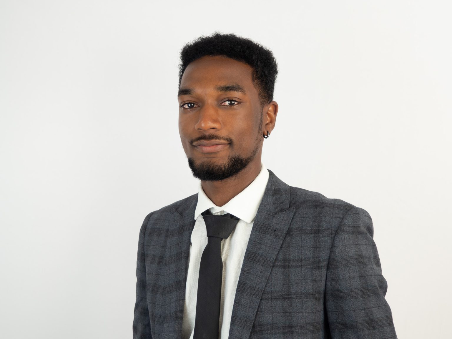 A professional headshot of a young Black man wearing a charcoal gray suit with a white dress shirt and black tie, smiling slightly at the camera against a light gray background.