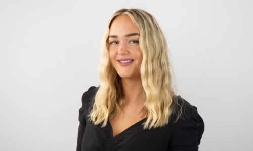 A smiling blonde woman wearing a black wrap-style top poses against a plain white background in what appears to be a professional portrait or headshot.