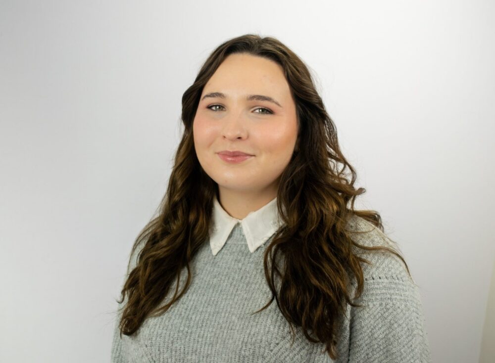 A woman with long wavy brown hair smiles at the camera, wearing a light blue knit sweater over a white collared shirt against a plain white background.