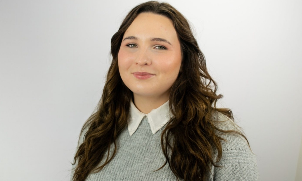 A woman with long wavy brown hair smiles at the camera, wearing a light blue knit sweater over a white collared shirt against a plain white background.