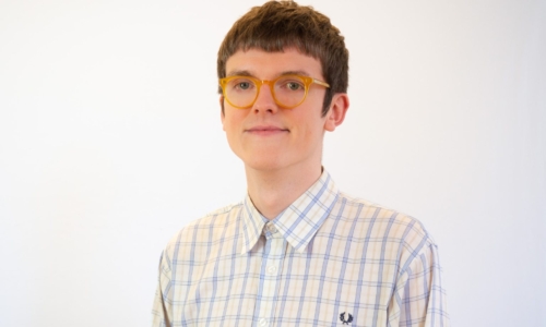 A person with short brown hair and yellow-framed glasses wears a light-colored plaid button-up shirt with blue stripes against a white background.