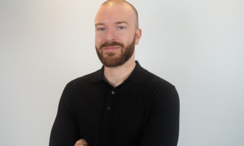 A man with a shaved head and brown beard wearing a black polo shirt stands with arms crossed against a white background, wearing a watch on his wrist.