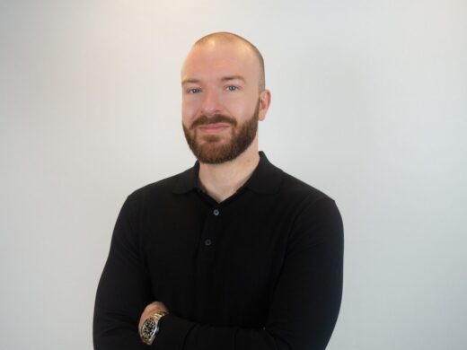 A man with a shaved head and brown beard wearing a black polo shirt stands with arms crossed against a white background, wearing a watch on his wrist.