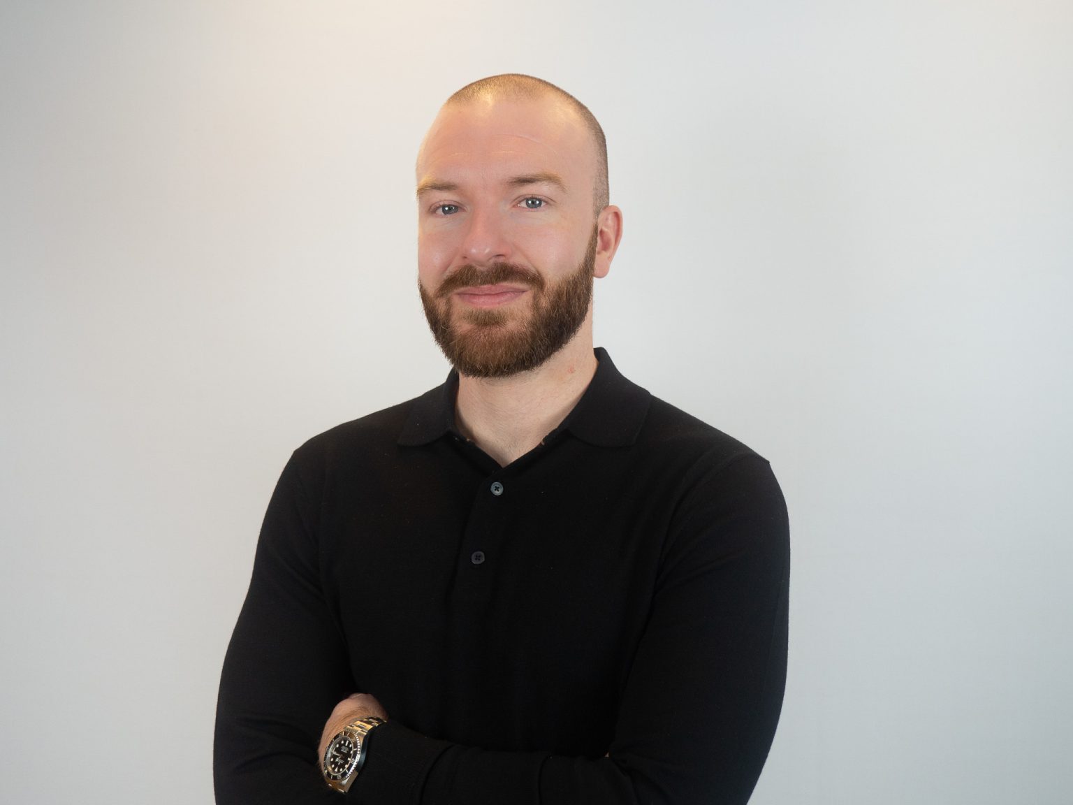 A man with a shaved head and brown beard wearing a black polo shirt stands with arms crossed against a white background, wearing a watch on his wrist.