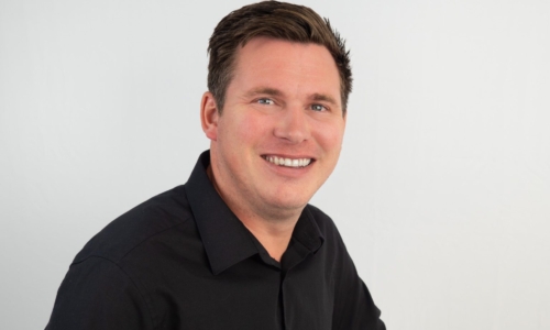 A smiling man with short brown hair wearing a black button-up shirt poses against a white background in what appears to be a professional headshot photograph.