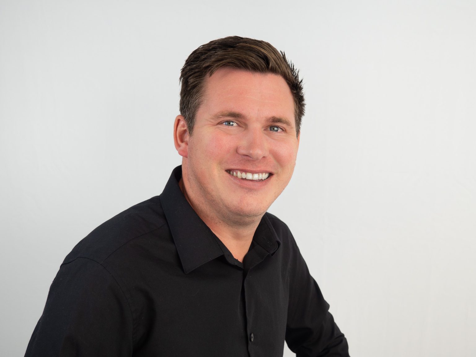 A smiling man with short brown hair wearing a black button-up shirt poses against a white background in what appears to be a professional headshot photograph.