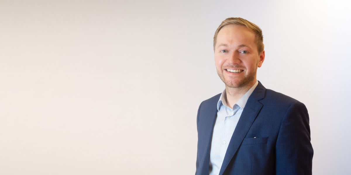 A smiling professional man with blonde hair and facial hair wearing a dark blue suit jacket and light blue striped dress shirt, photographed against a neutral background in what appears to be a corporate headshot or business portrait.