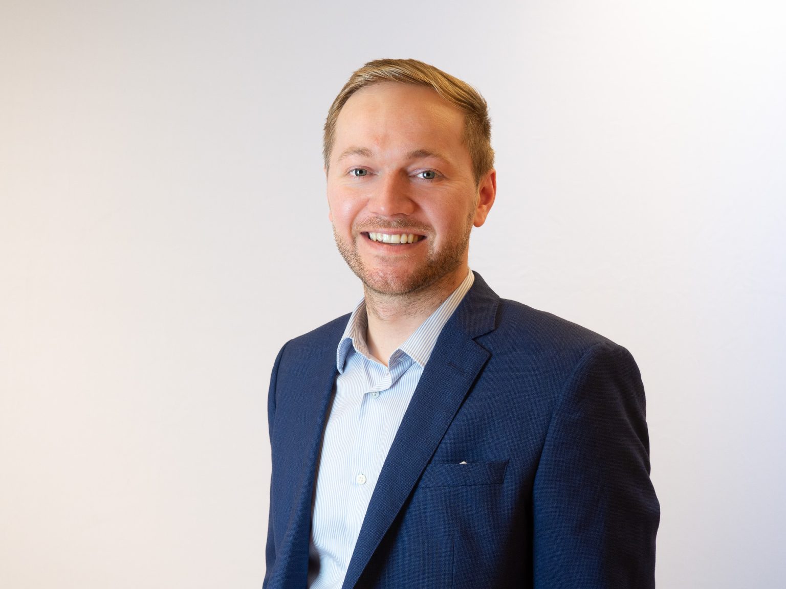 A smiling man with blonde hair and facial stubble wearing a navy blue blazer over a light blue striped dress shirt, photographed against a plain white background in a professional headshot style.