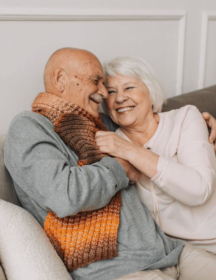 An elderly couple embraces warmly while sitting on a couch, both smiling joyfully with their foreheads touching in an intimate, loving moment.