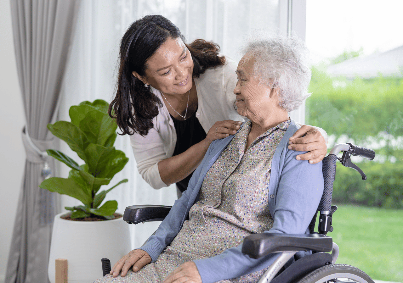 A caregiver gently places her hands on the shoulders of an elderly woman in a wheelchair, both smiling warmly in a bright room with large windows overlooking greenery.