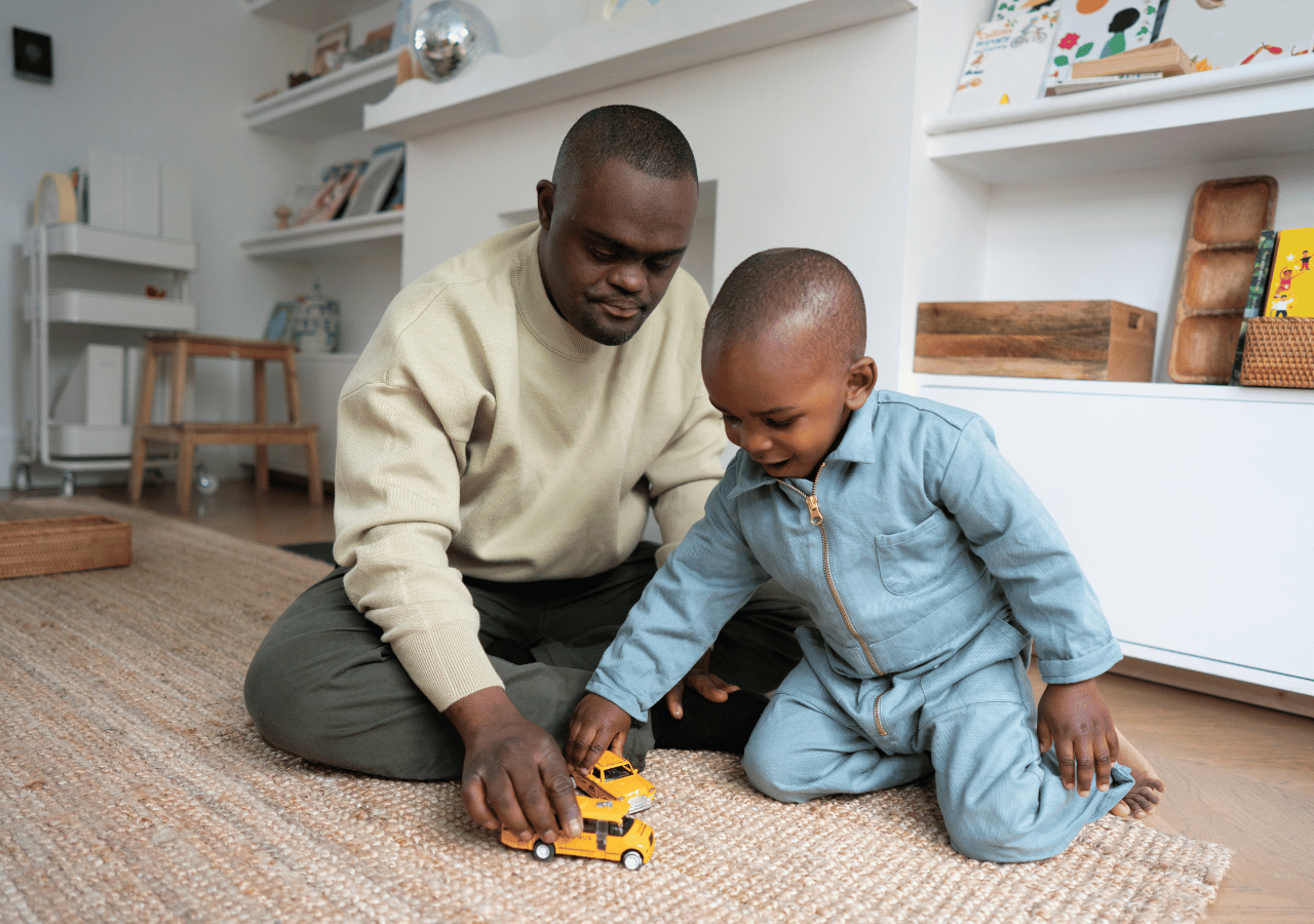 A father and young child sit on a textured rug playing with a yellow toy truck in a modern living room with white built-in shelving filled with books and decorative items.
