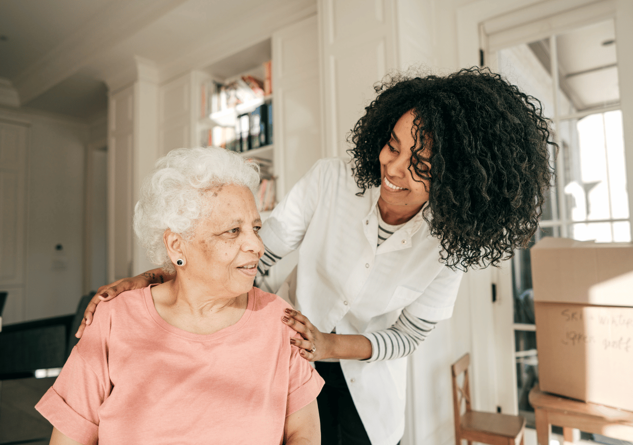 A smiling healthcare worker with curly dark hair wearing a white coat places a caring hand on the shoulder of an elderly woman with white hair wearing a pink top, both appearing happy in what looks like a bright, modern home or care facility.