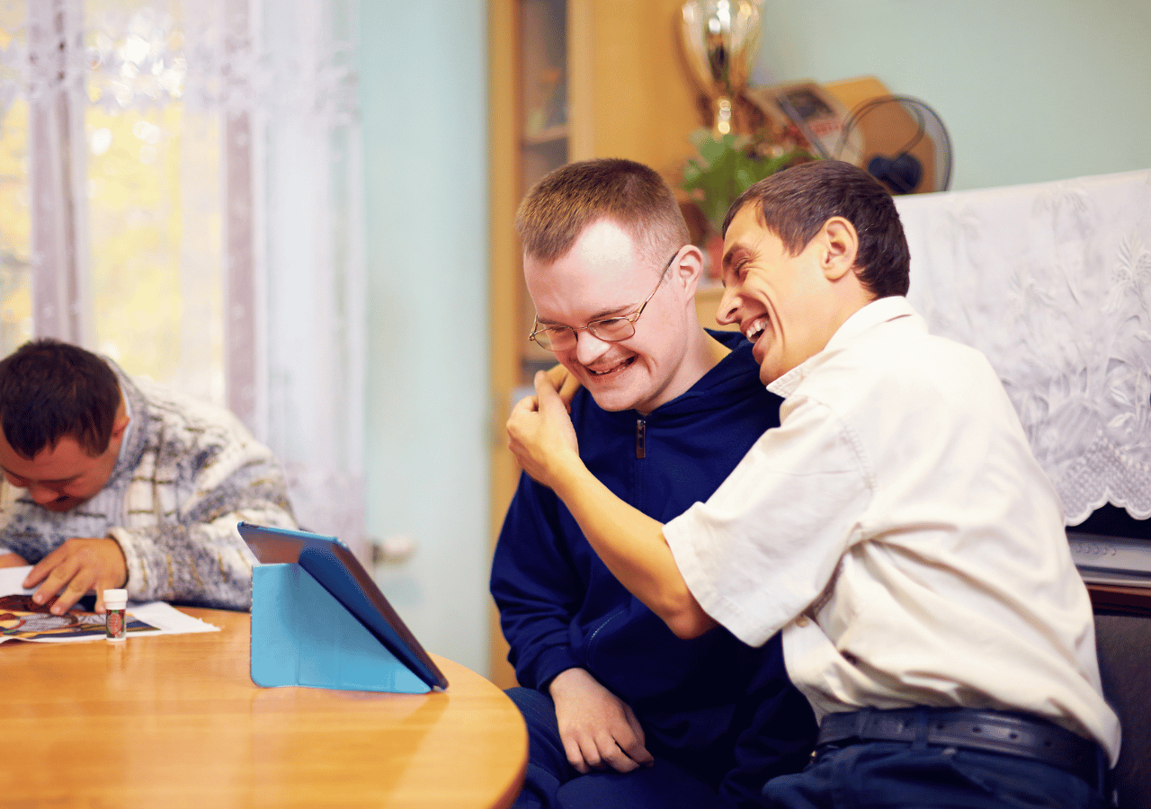 Two men with disabilities sit together at a table, smiling and laughing while one has his arm around the other's shoulder in a friendly embrace. A third person works at the table in the background, with a tablet visible on the wooden surface in what appears to be a supportive community or care setting.