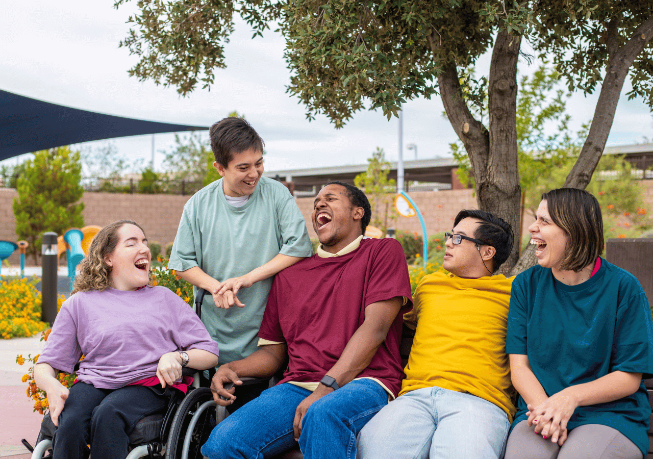 A diverse group of five young people with disabilities laughing and socializing together outdoors under a shade structure, with one person in a wheelchair and colorful flowers visible in the background.