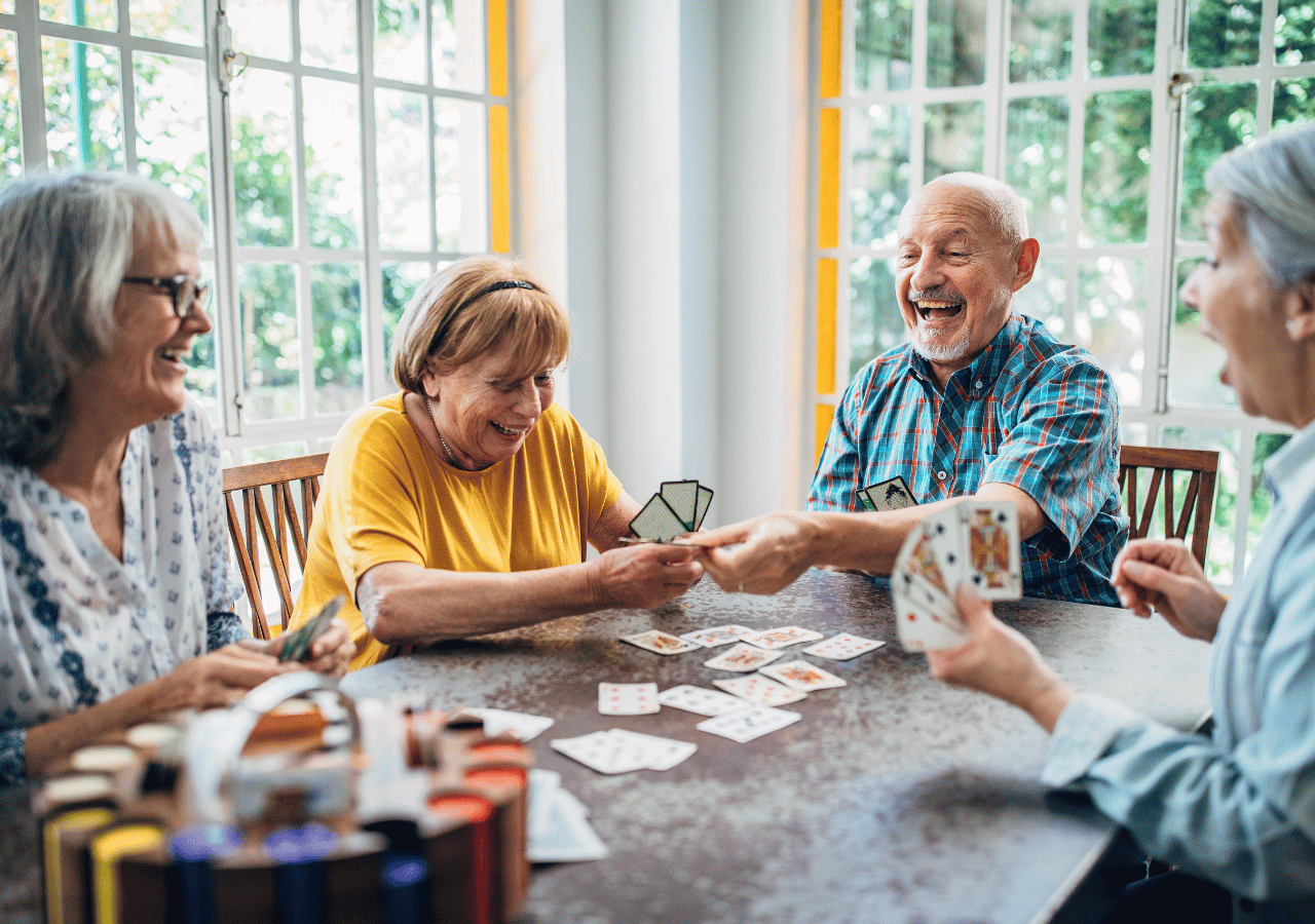 Four older adults sit around a table playing cards in a bright, sunlit room with large windows. They appear to be enjoying themselves, with one man laughing heartily as they engage in their card game together.