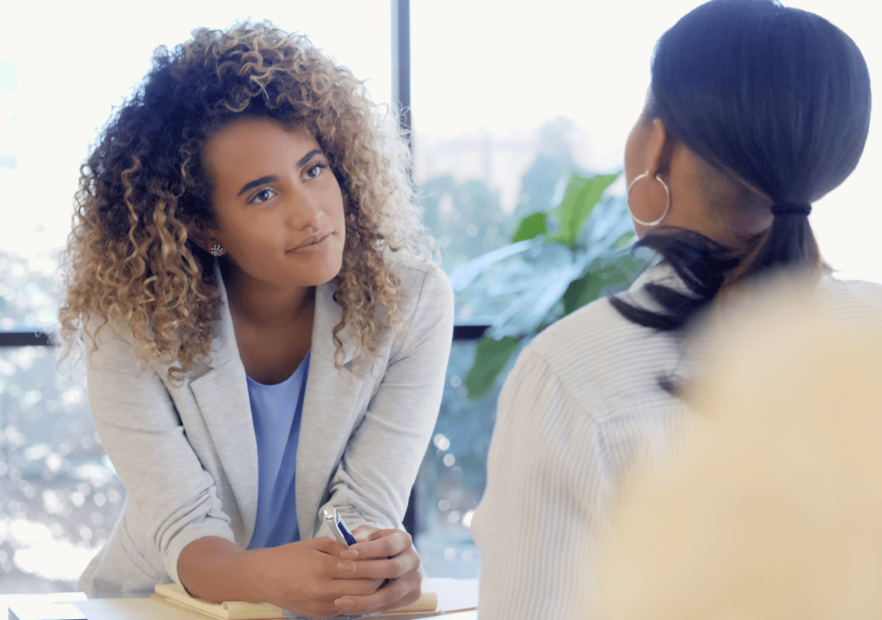 Two women are having a professional conversation in a bright office setting, with one woman in a light blazer facing another woman whose back is to the camera, suggesting a business meeting or interview scenario.