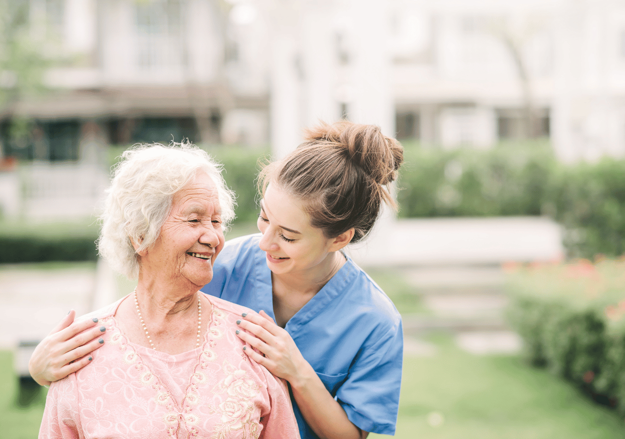 A young healthcare worker in blue scrubs embraces an elderly woman with white hair and a pink cardigan, both smiling warmly while standing outdoors near a care facility.